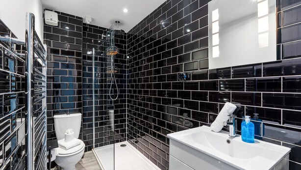 A modern bathroom with black tiled walls, a white toilet, a glass-enclosed shower, and a white sink with a mirror above it. - Seaside Haven in Brighton (Brighton)