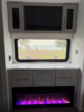 A compact RV interior with a flat-screen TV mounted above a window, a fireplace below, and a small storage area with drawers. - The Coastal Caravan (Alberton)