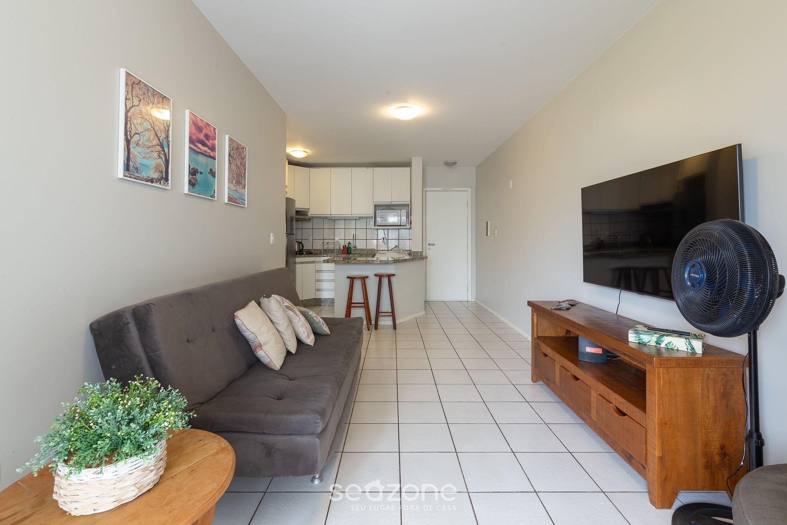 A living room with a sofa, a TV, a fan, and a kitchen in the background.