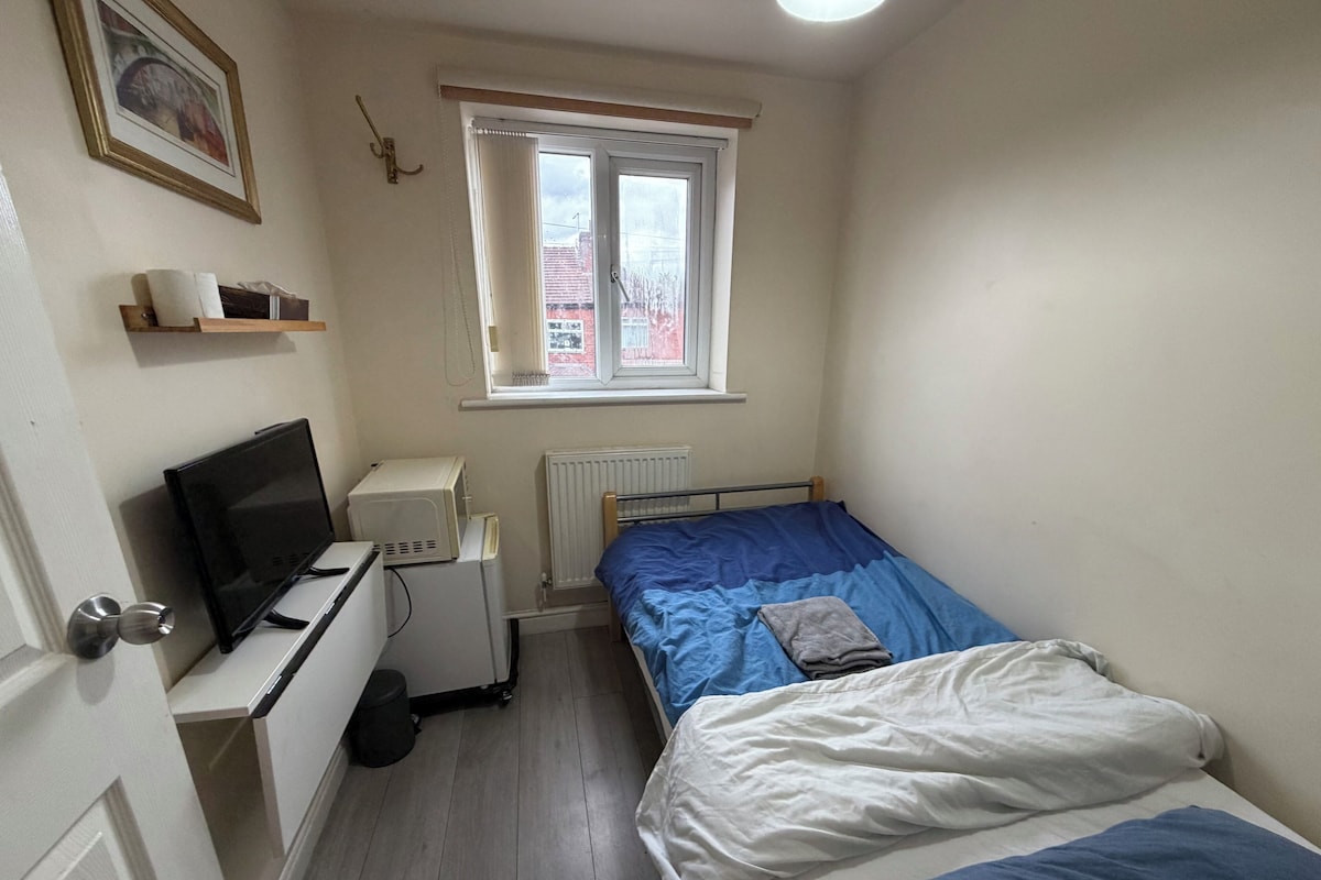 A small, tidy room with a single bed, a desk with a computer, a radiator, and a window with a view of buildings.