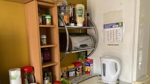A kitchen with a wooden shelf holding condiments, a basket of fruit, and a kettle on the counter. - Hotel Jacks Court (Ashton-under-Lyne)