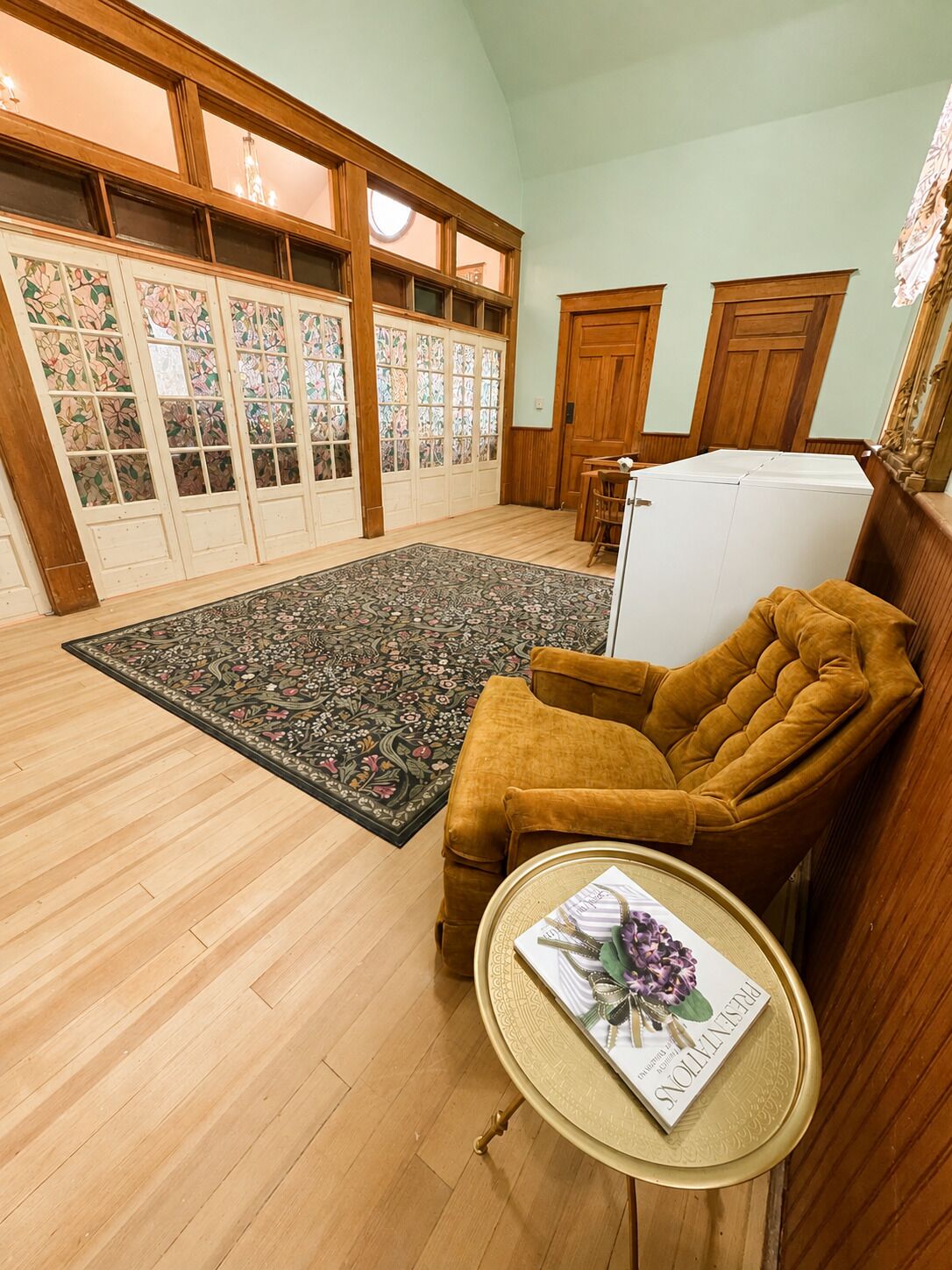 Een kamer met een houten vloer, een kamerbreed tapijt met patroon, een bruine fauteuil, een klein rond tafeltje met een boek, en een wit apparaat.