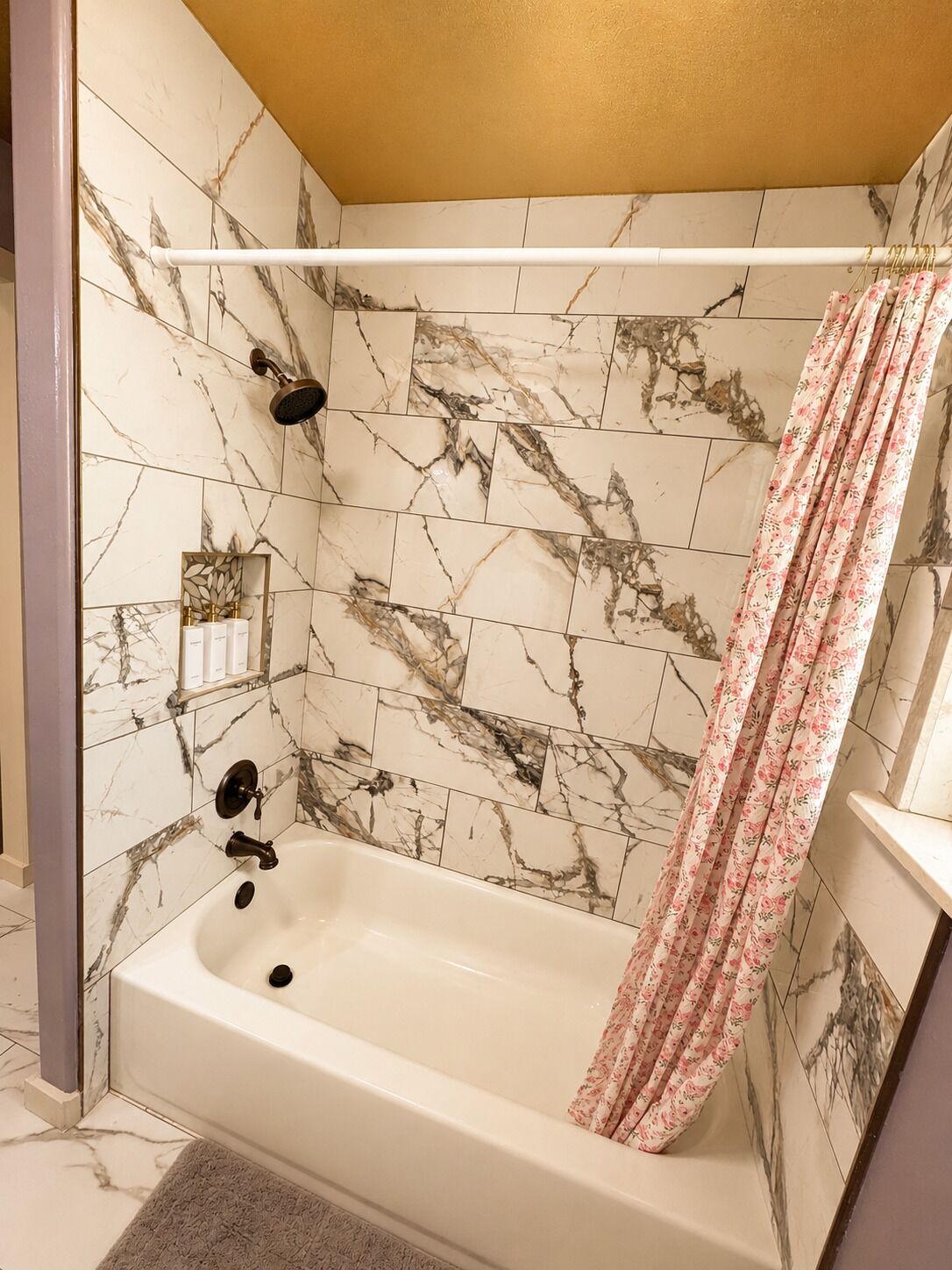 A bathroom with a marble-tiled wall, a bathtub, a showerhead, and a floral shower curtain.