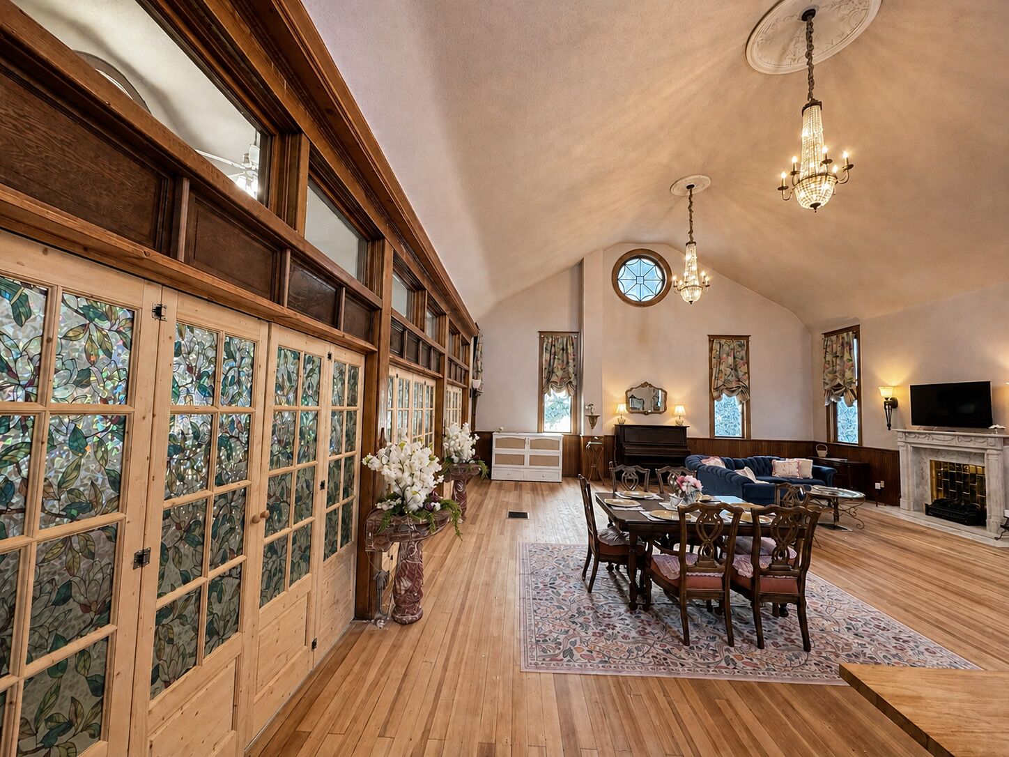 A spacious living area with a wooden floor, a dining table with chairs, a fireplace, and a large window with stained glass.
