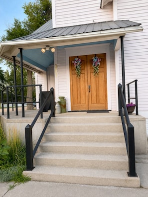 A house with a front porch, double wooden doors, and a staircase leading to the entrance. - Charming 1907 Chapel - Luxurious Retreat Near Fort Morgan & Prewitt Reservoir (Hillrose)
