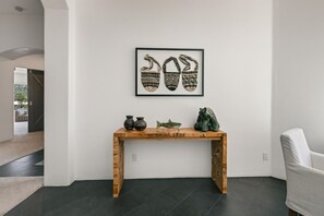 Ein schlicht eingerichteter Raum mit dunklem Boden, einer weißen Wand, einem gerahmten Kunstwerk über einer Holzkonsole und einem weißen Stuhl zur Rechten.
