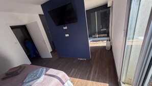 Une chambre d’hôtel avec un lit, un téléviseur à écran plat fixé au mur, une porte-fenêtre coulissante donnant sur un balcon et une valise blanche posée sur le sol.