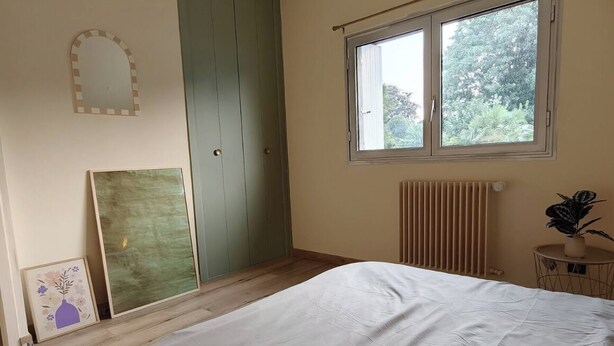 Une chambre à coucher avec un lit, une fenêtre, un radiateur, une armoire verte et un miroir au mur.