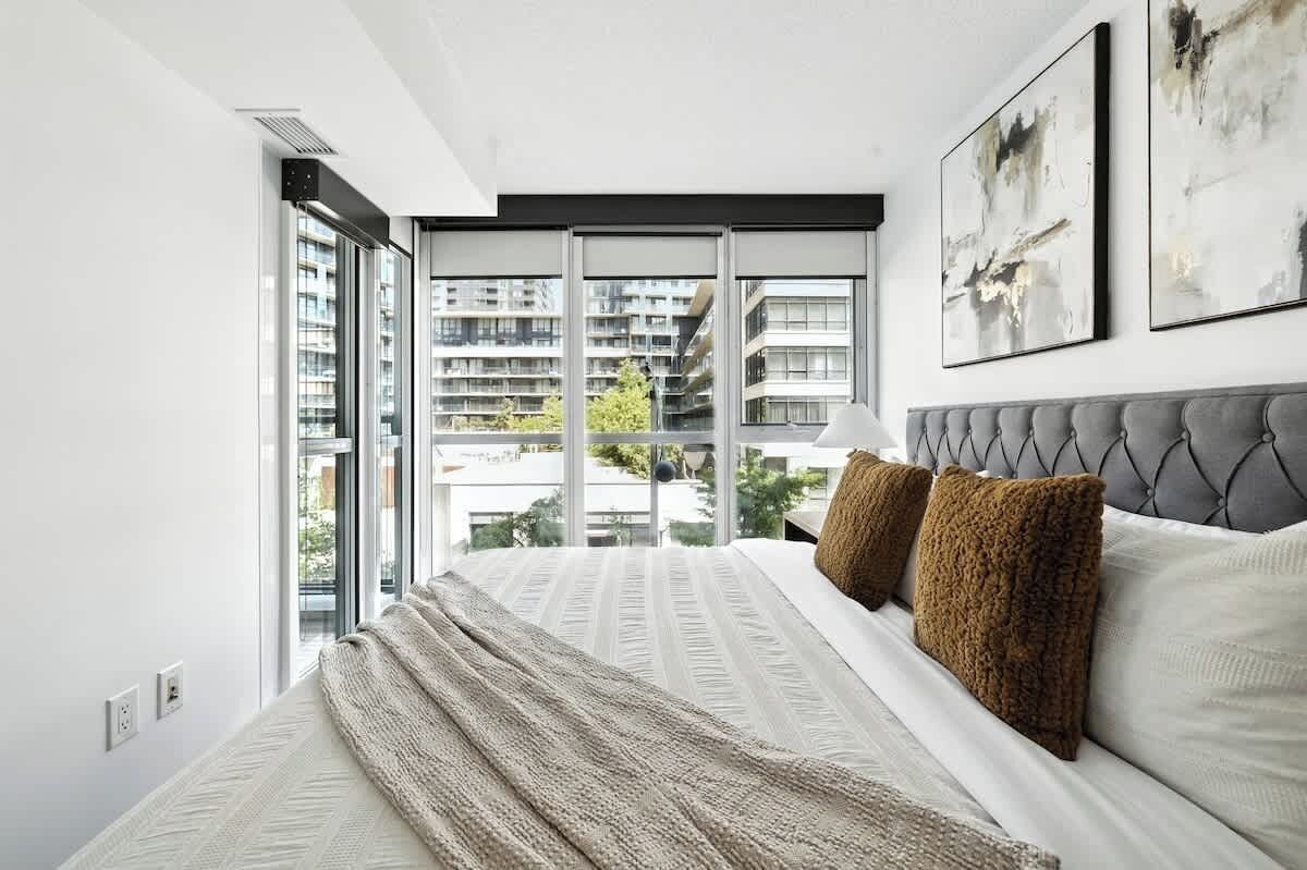 A modern bedroom with a large window, a tufted headboard, and abstract wall art.
