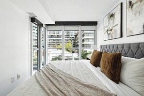 A modern bedroom with a large window, a tufted headboard, and abstract wall art. - CityPlace Skyline Retreat Sleeps 5 w Patio! (Toronto)