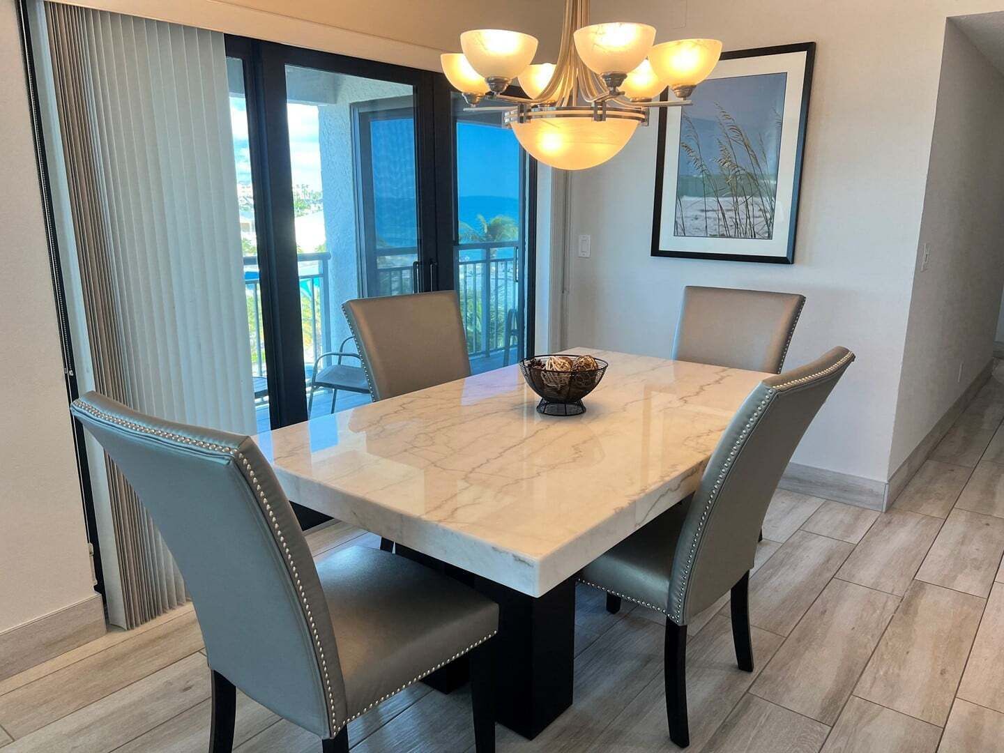 A dining area with a marble table, four chairs, a chandelier, and a view of the outdoors.