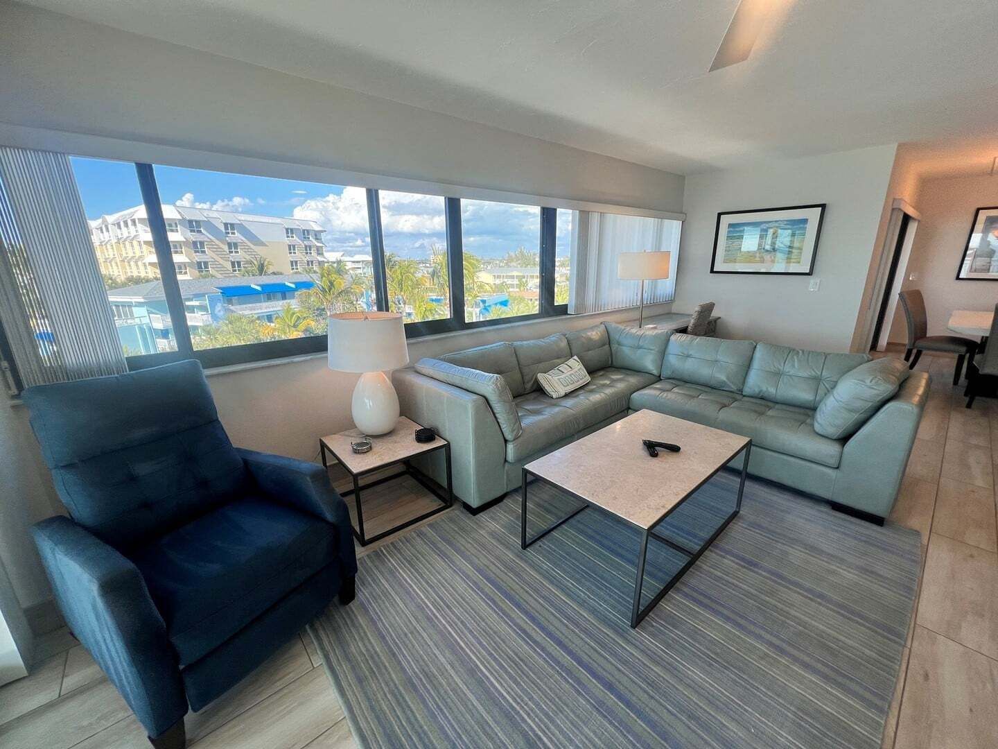 A modern living room with a sectional sofa, a coffee table, a side chair, and a lamp. Large windows provide a view of buildings outside.