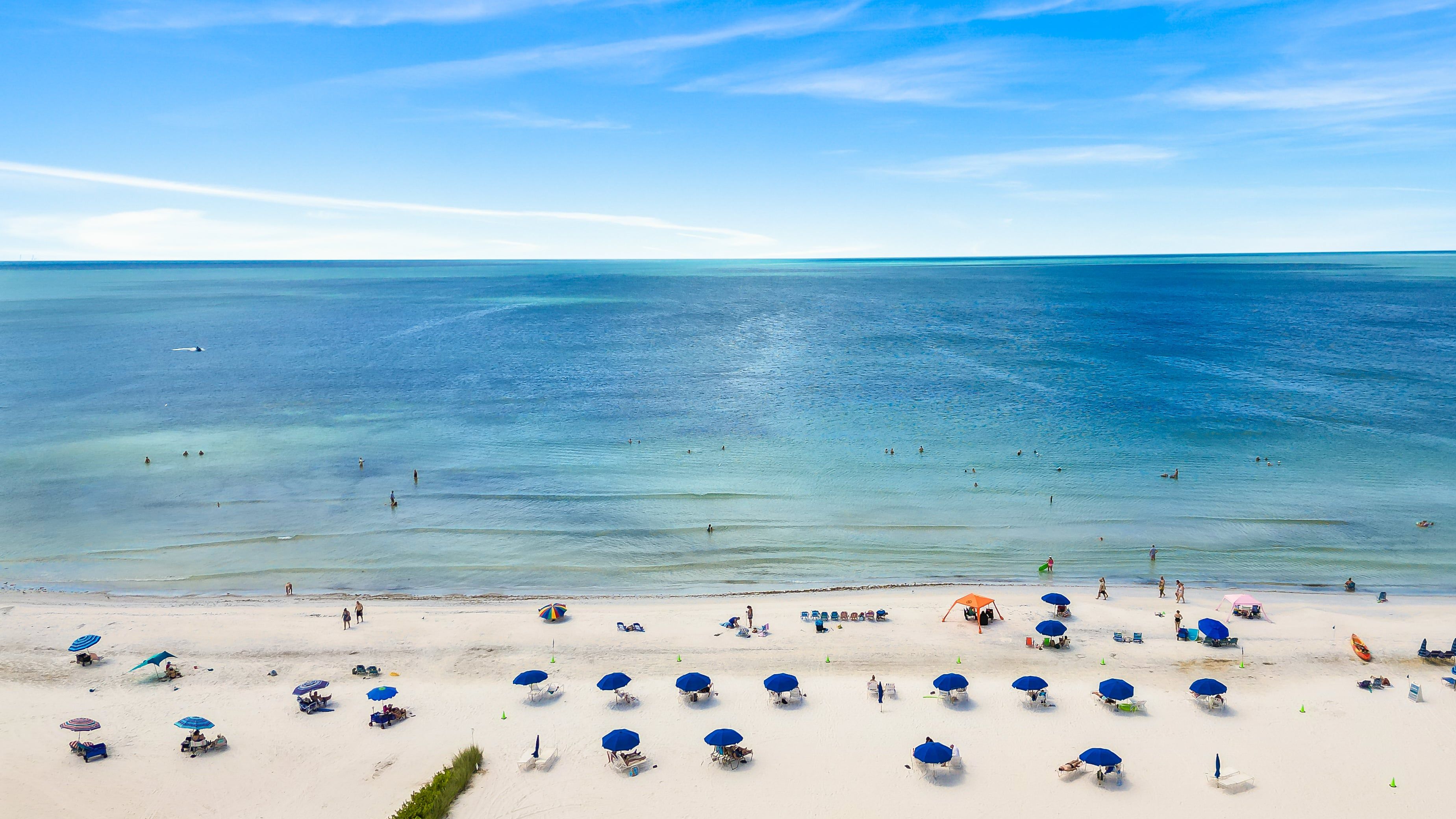 Een strand met helderblauw water, mensen die zwemmen en tal van strandparasols en ligstoelen.