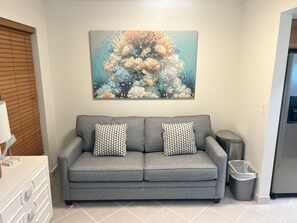 A grey sofa with patterned cushions, a wall-mounted painting of coral and fish, a trash bin, and a water dispenser. - Siesta Sands - Unit 412 (Siesta Key)