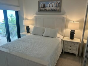 A bedroom with a large bed, two bedside tables with lamps, a painting above the bed, and a balcony with a view. - Siesta Sands - Unit 412 (Siesta Key)
