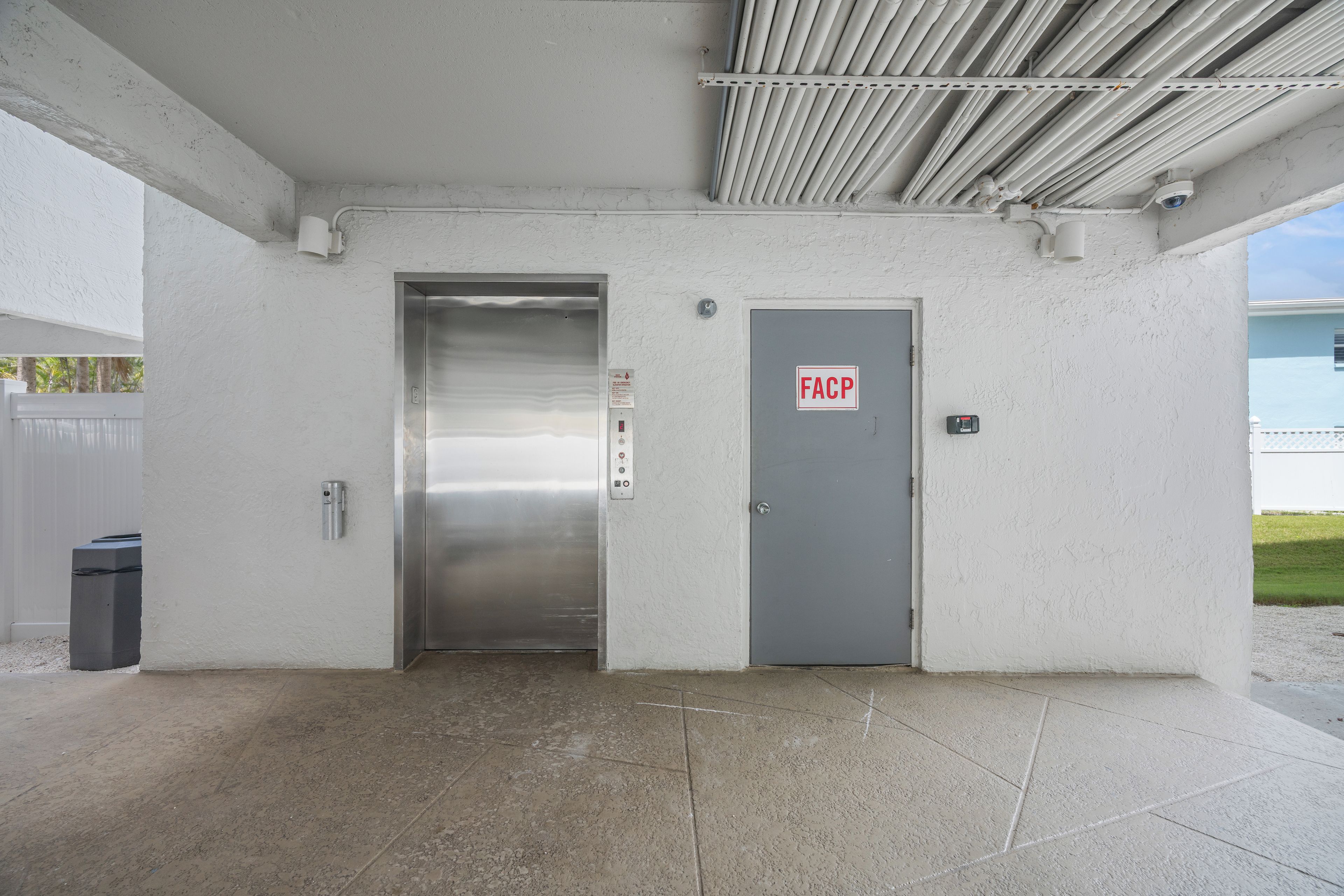 En bygningseingang med elevator i metal, en dør med teksten 'FACP' og et overvågningskamera.