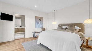 Ein modernes Schlafzimmer mit einem großen Bett, einem Nachttisch, einem Fernseher und einem durch eine offene Tür einsehbaren Badezimmer.