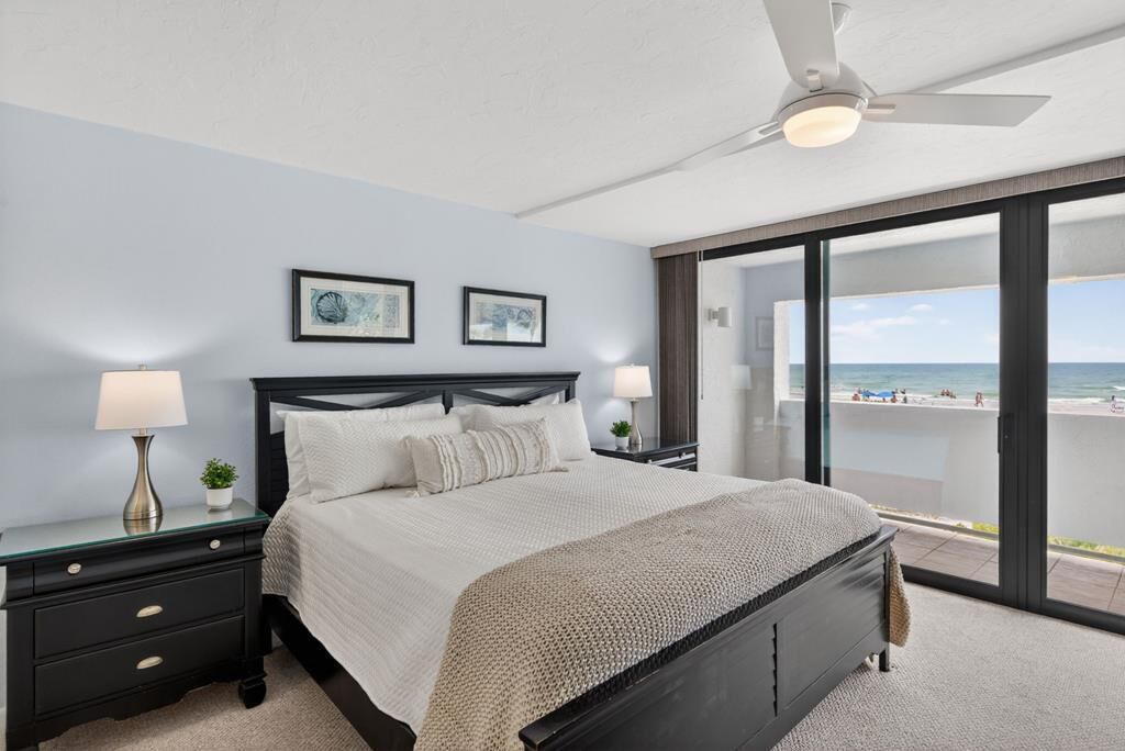 Ein Schlafzimmer mit einem Bett, zwei Nachttischen, einem Deckenventilator und Blick auf den Strand.
