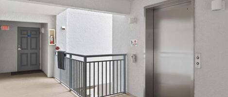Un pasillo con barandal de metal, un ascensor y una puerta con letrero de salida de emergencia.
