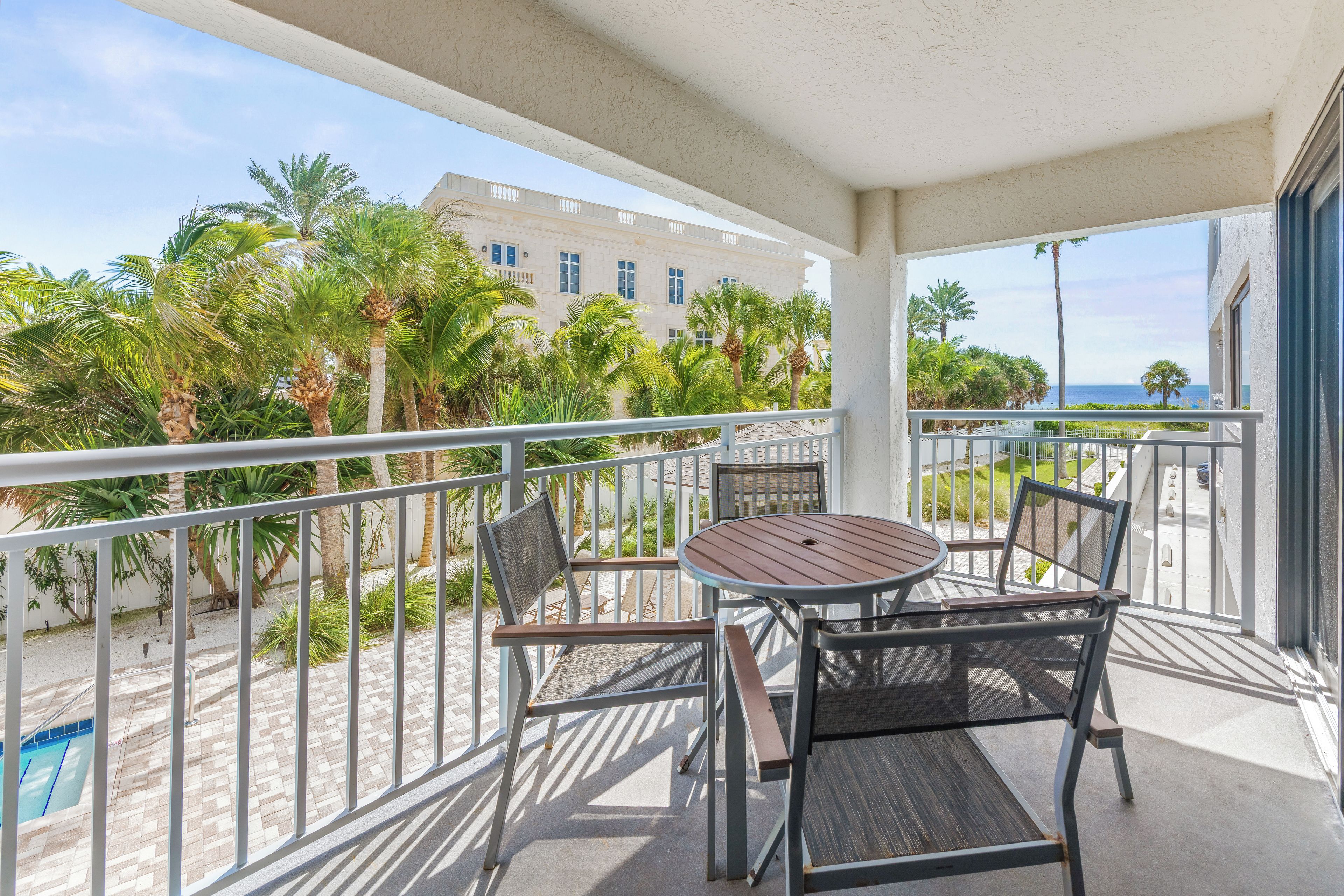 A balcony with a table and chairs, overlooking a pool and palm trees, with a building in the background.