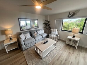 A living room with a grey sofa, a white coffee table, a striped armchair, a side table with a lamp, a large mirror, and a ceiling fan. - Siesta Sands Unit 309 (Siesta Key)