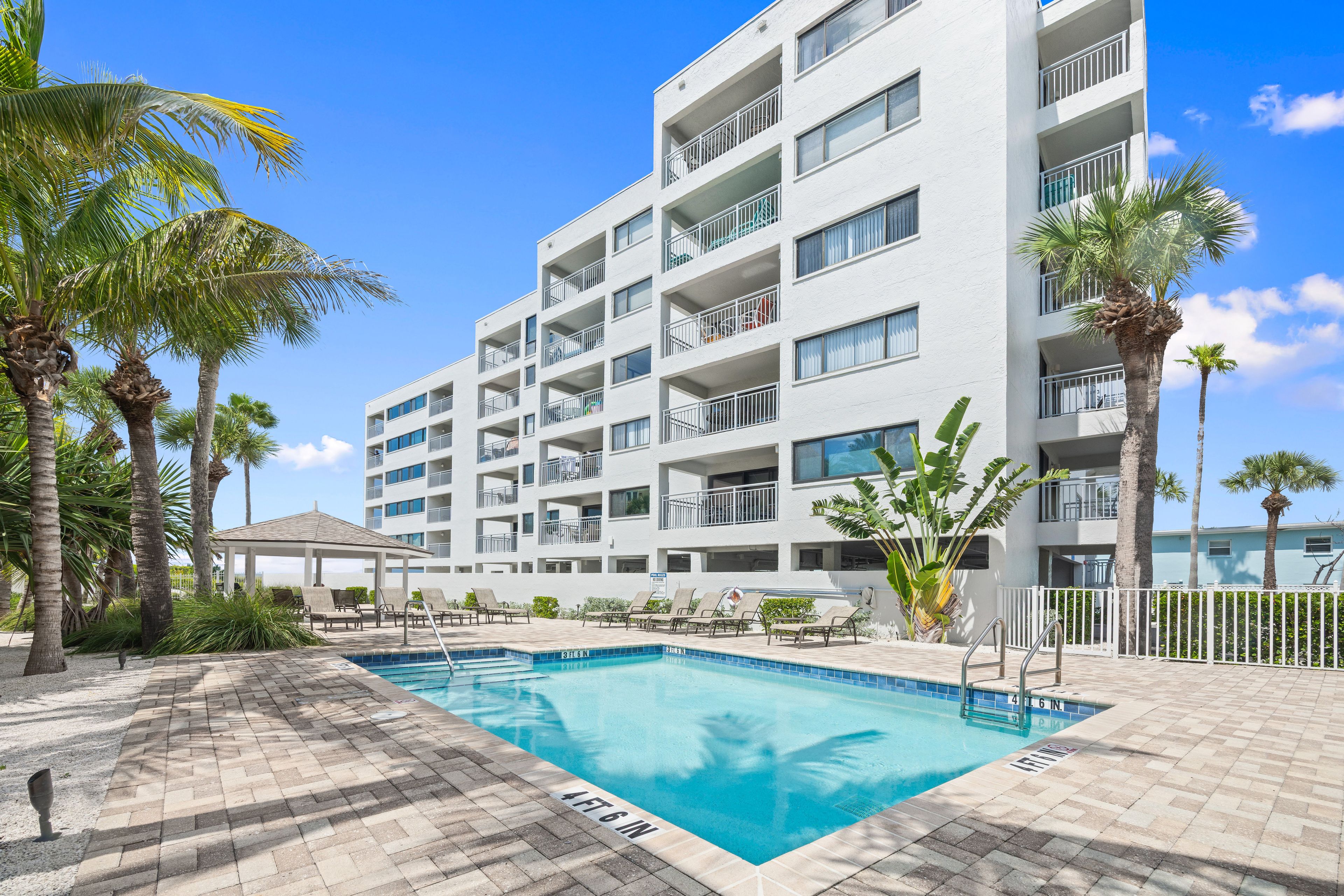 A multi-story hotel with a swimming pool, palm trees, and a covered seating area.