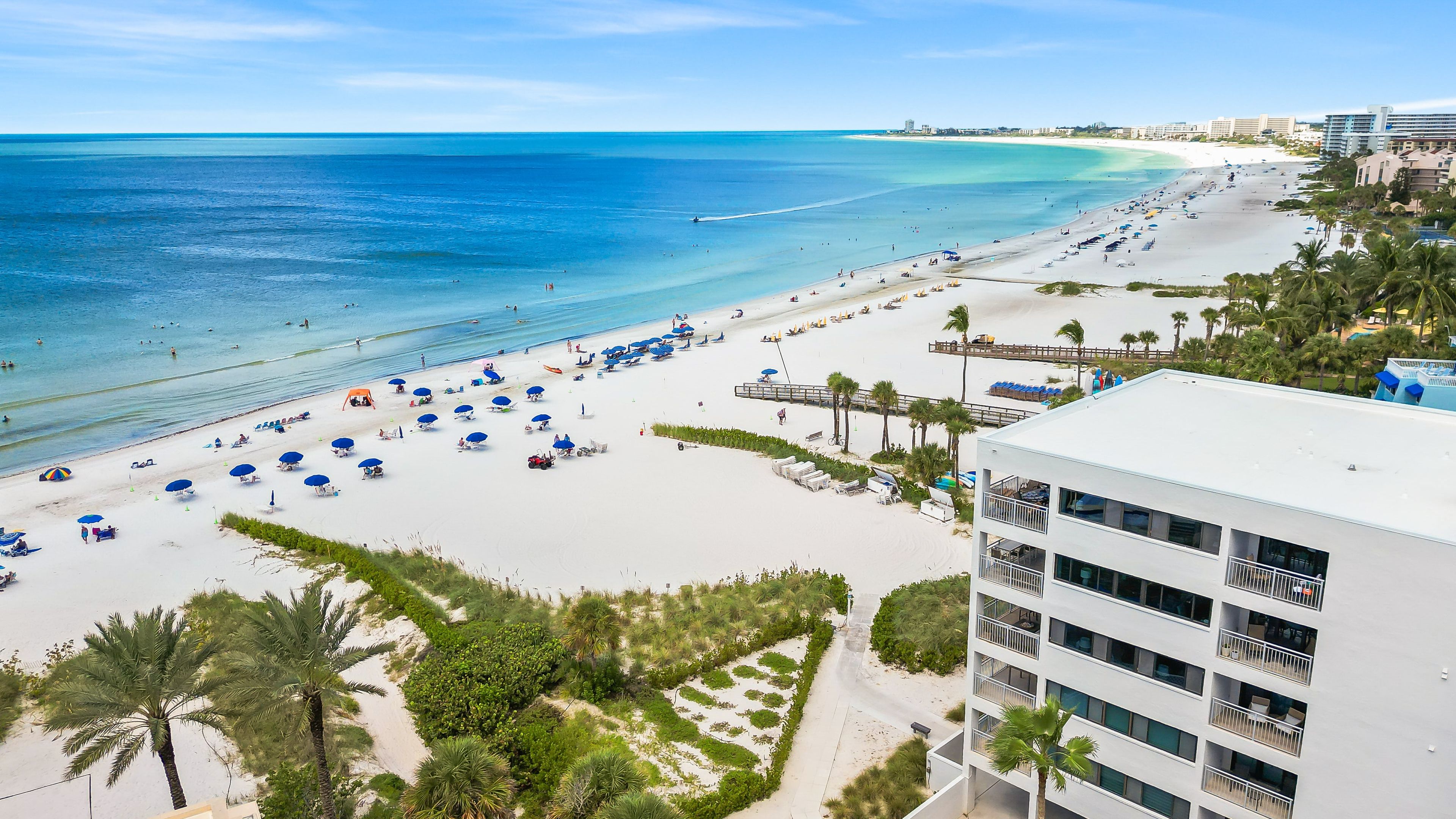 A beach with white sand, clear blue water, and a modern building with balconies.