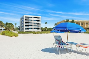 Ein Strand mit weißem Sand, einem blauen Sonnenschirm und einem mehrstöckigen Gebäude im Hintergrund.