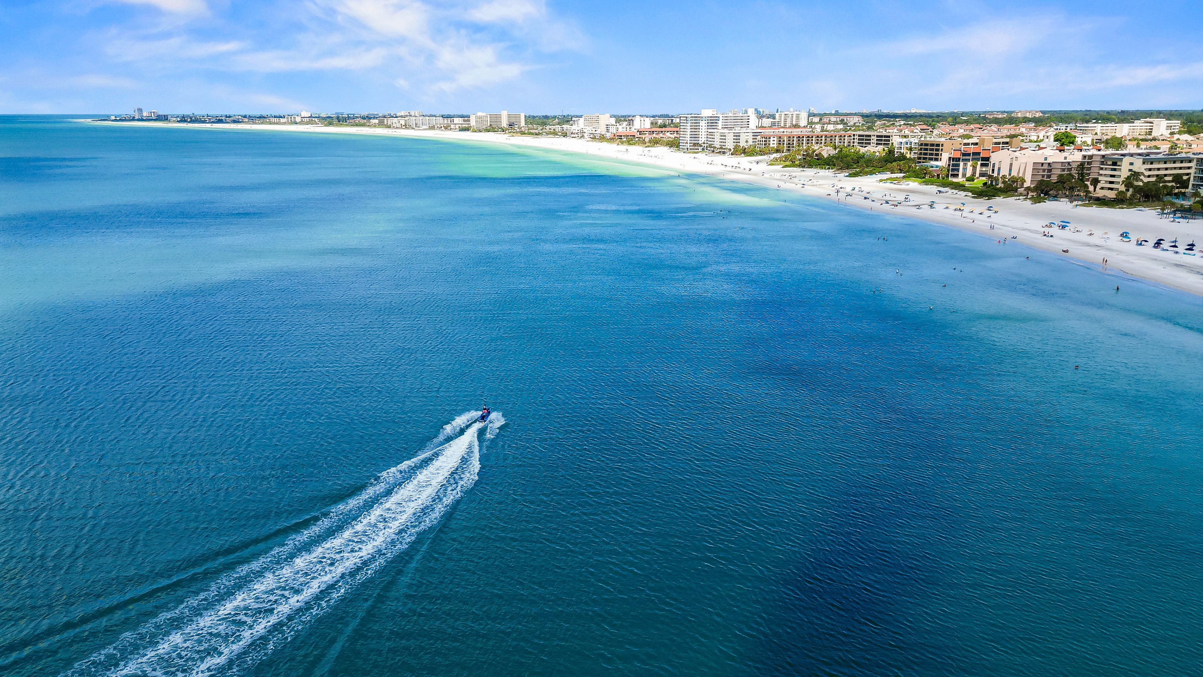 An aerial view of a coastal area with a beach, buildings, and a boat creating a wake in the water.