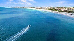Vue aérienne d’une zone côtière avec une plage, un yacht laissant une vague derrière lui et une vue sur la ville en arrière-plan.