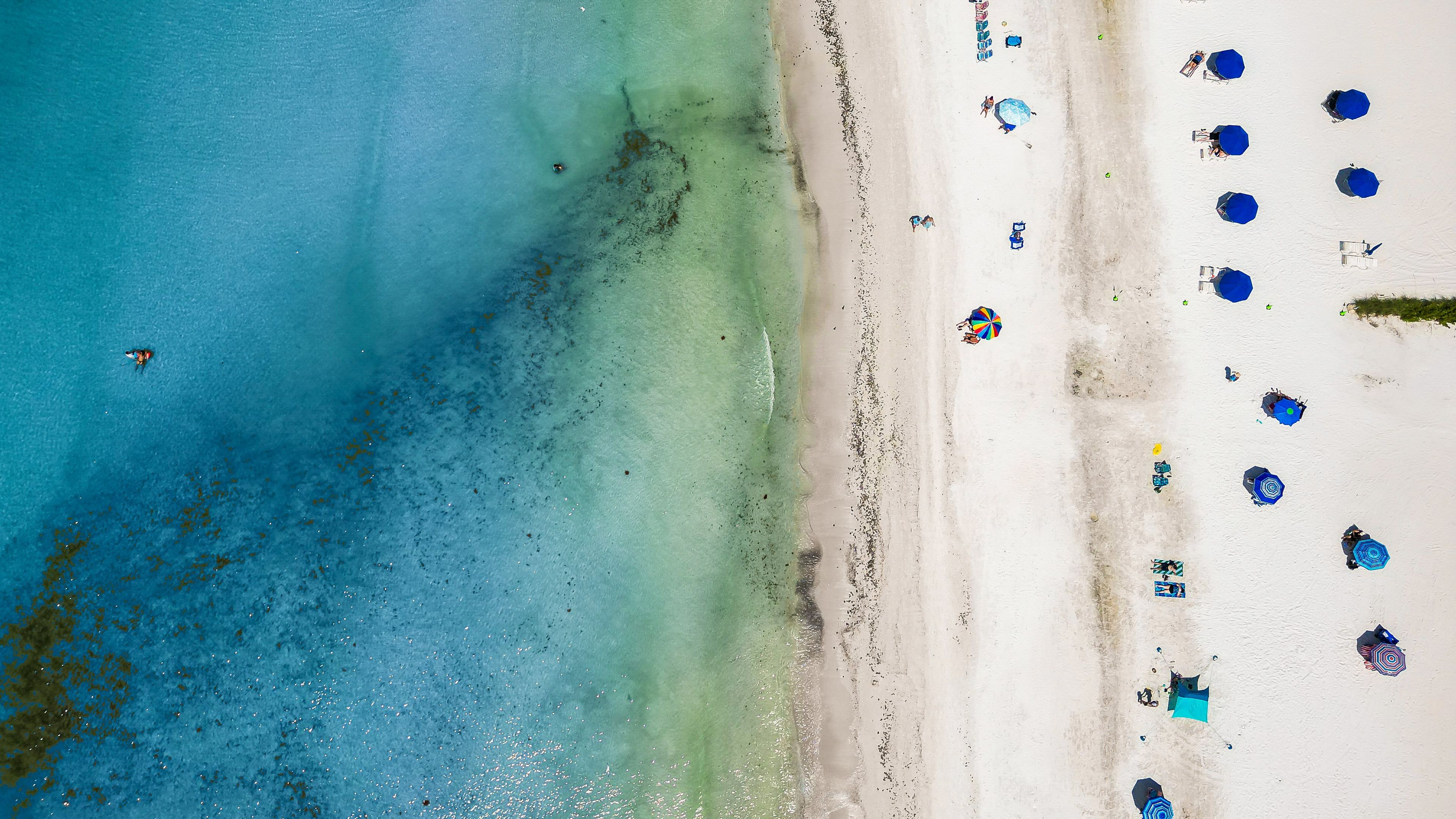 Eine Vogelperspektive zeigt einen Strand mit klarem türkisblauem Wasser und weißem Sand, gesäumt von mehreren blauen Sonnenschirmen.