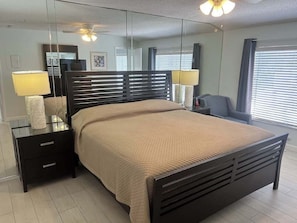 A modern bedroom with a large bed, bedside lamps, a nightstand, a mirror, and a ceiling fan. - Siesta Sands Unit 205 (Siesta Key)