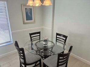 A dining area with a round glass table surrounded by four chairs, a framed picture on the wall, and a window with blinds. - Siesta Sands Unit 205 (Siesta Key)