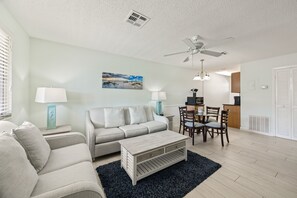 A living room with a sofa, a coffee table, a dining table with chairs, a ceiling fan, and a painting on the wall. - Siesta Sands - Unit 206 (Siesta Key)