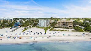 A beach with multiple umbrellas and lounge chairs, a clear sky, and buildings in the background. - Siesta Sands - Unit 206 (Siesta Key)