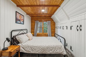 Un dormitorio con techo de madera, cama con ropa de cama floral, mesita de noche, lámpara, ventana con cortinas y armario empotrado.