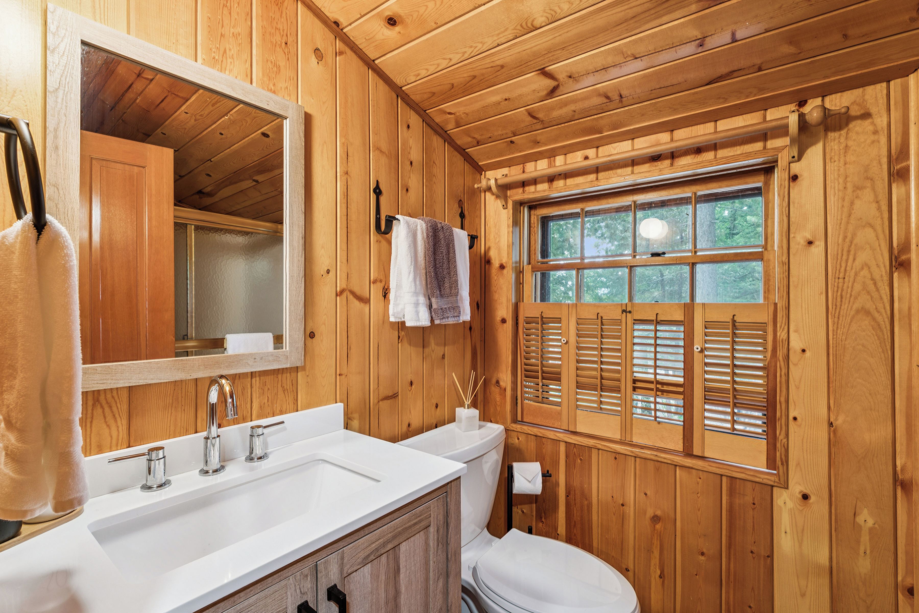 Une salle de bain rustique, aménagée dans une cabane en bois, avec un grand miroir, un lavabo, des toilettes et une fenêtre équipée de volets.