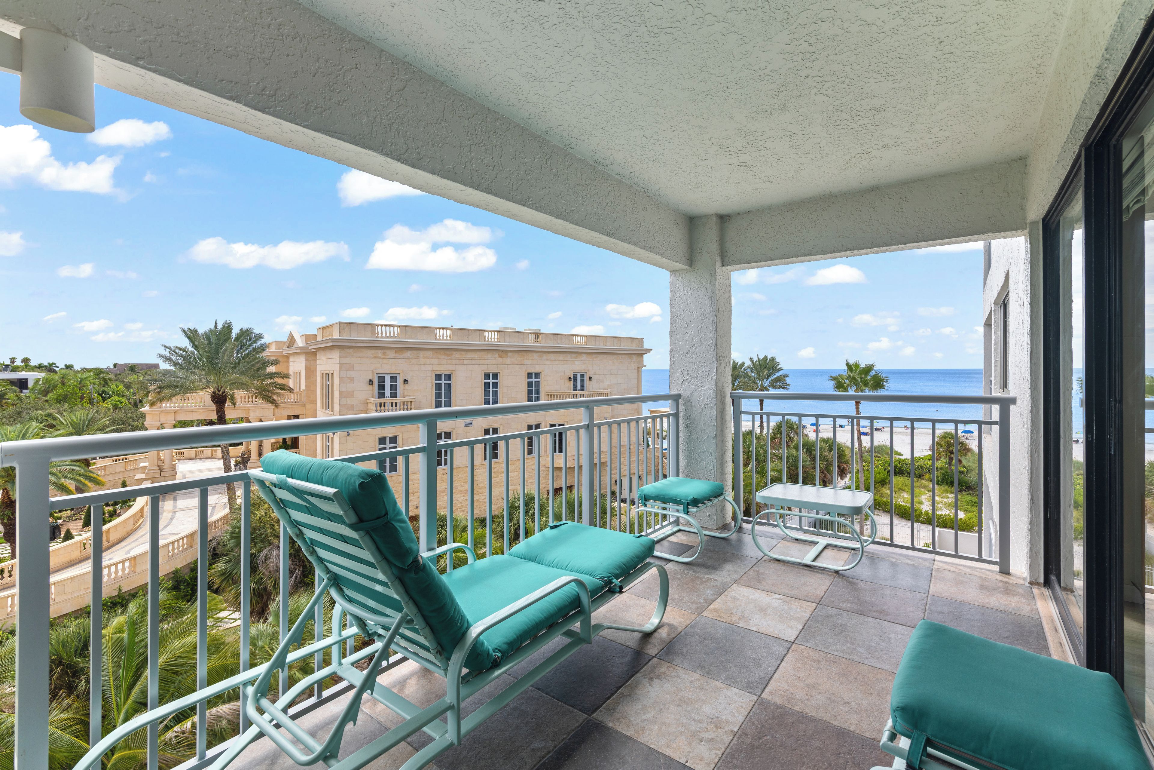 A balcony with two green cushioned chairs and a small table, overlooking a beach and a building.