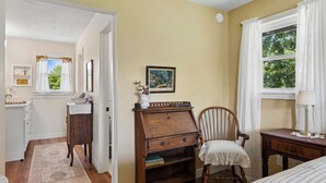 Una habitación con escritorio de madera, una silla, una cama, un ventanal con cortinas y un cuadro en la pared.