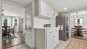 Una cocina moderna con armarios blancos, un refrigerador de acero inoxidable y una zona de comedor con una mesa y sillas de madera.