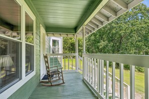 A covered porch with a rocking chair, a window, and a view of a green landscape.