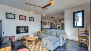 A modern living room with a grey sofa, a wooden coffee table, and a television. There are abstract art pieces on the wall and a window with blinds. - Ti Kaz Komela - Vue montagnes - Terrasse (Le Tampon)