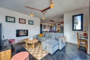 A modern living room with a grey sofa, a wooden coffee table, and a television. There are abstract art pieces on the wall and a window with blinds. - Ti Kaz Komela - Vue montagnes - Terrasse (Le Tampon)