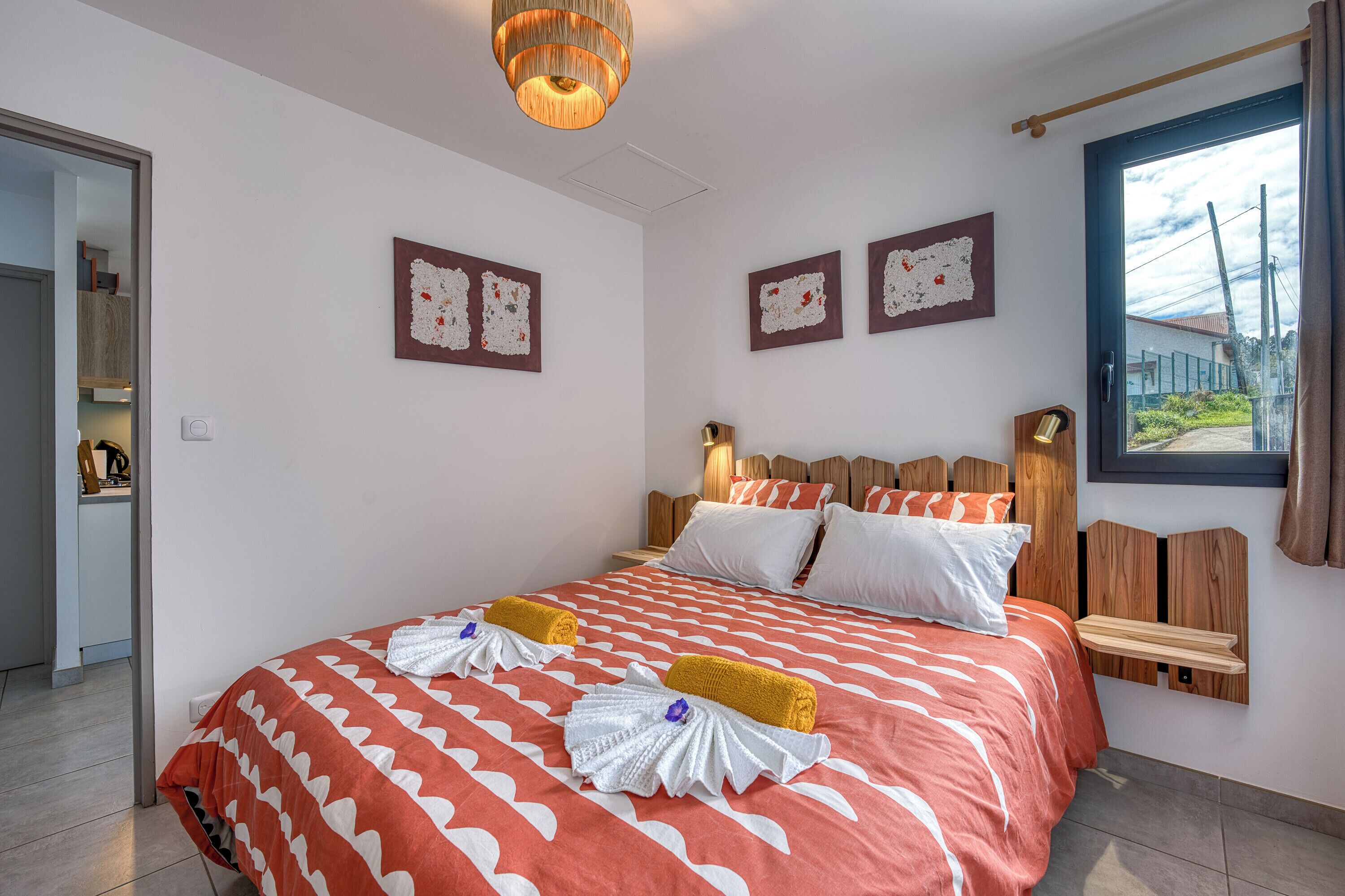 A hotel room with a large bed, wooden headboard, two wall-mounted picture frames, a window with curtains, and a door leading to a bathroom.