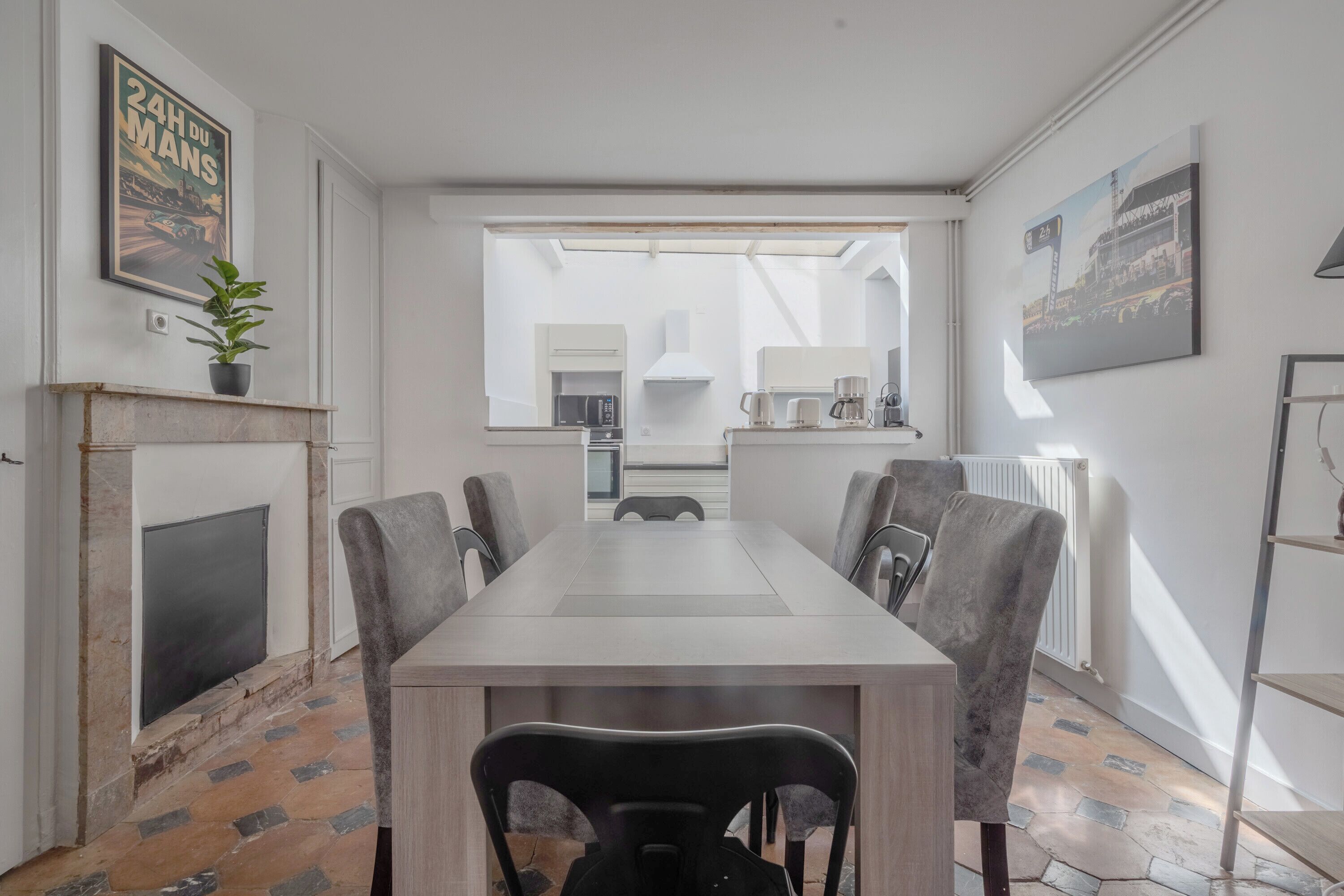 A modern dining room with a long table, grey upholstered chairs, a fireplace, and a large window with a view of the kitchen.