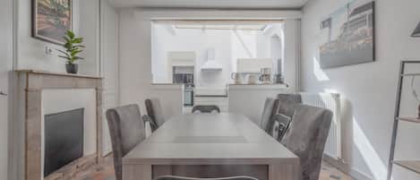 A modern dining room with a long table, grey upholstered chairs, a fireplace, and a large window with a view of the kitchen.