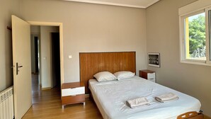 Ein Schlafzimmer mit einem hölzernen Bett, einem Nachttisch, einer Leselampe und einem Fenster mit Blick auf Grünflächen.