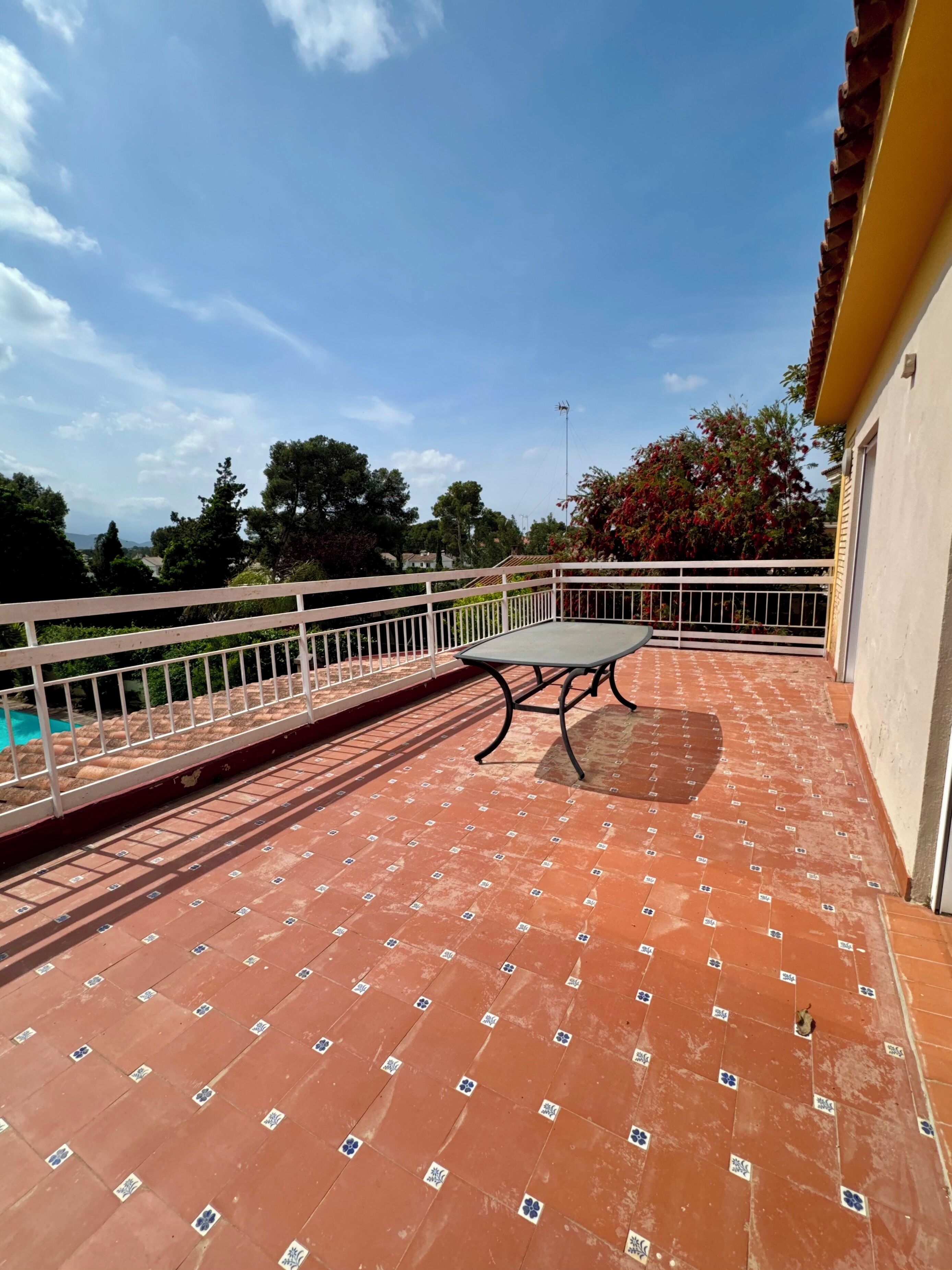 Una terraza con mesa y sillas, rodeada por una barandilla blanca, y vistas a árboles y un paisaje lejano.