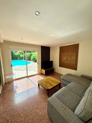 Un salon avec un canapé gris, une table basse en bois et un téléviseur posé sur un meuble. Une porte-fenêtre coulissante donne accès à un espace piscine.