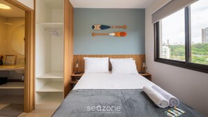 Quarto de hotel com cama, guarda-roupa de madeira, mesa de cabeceira e vista da cidade através de uma janela grande.